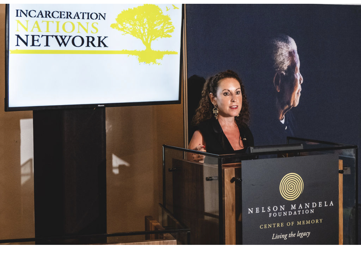 Dr. Baz Dreisinger speaks at a podium at the Nelsmon Mandela Foundation with a background slide of Incarceration Nations Network.