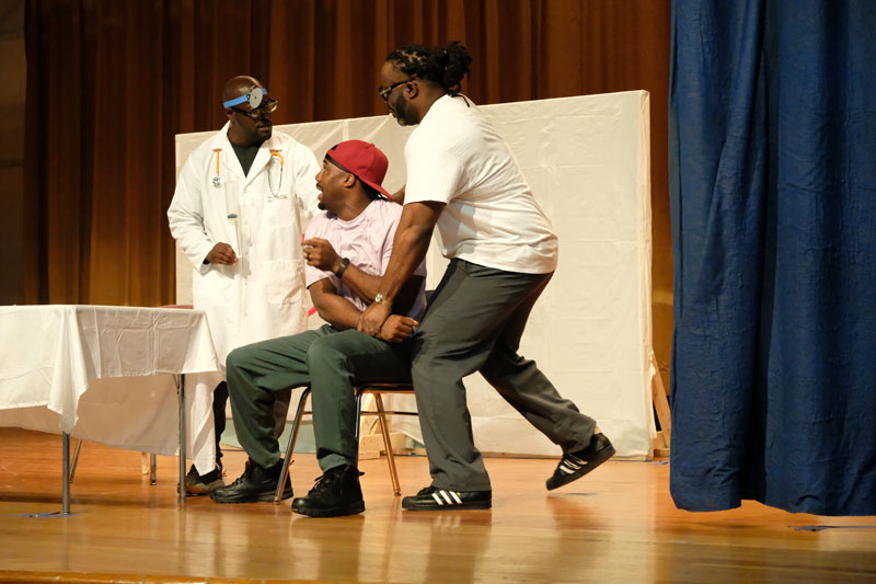 A scene from A Passing Life. A man in a chair fearfully looks up at a doctor in a white coat.