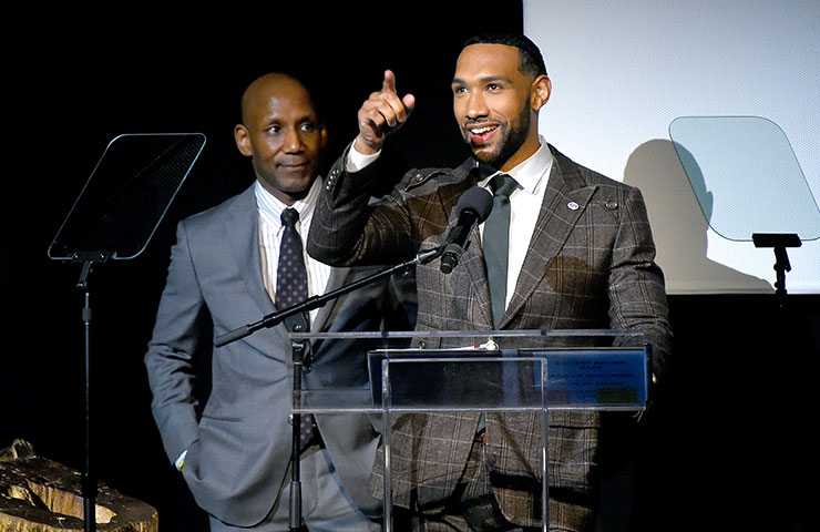 Craig Steven Wilder stands behind Dyjuan Tatro as he speaks on stage and points up.