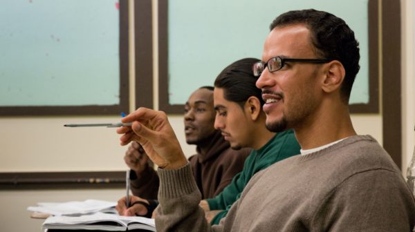 BPI student speaking in class, gesturing with his pen, while other students write notes in the background.