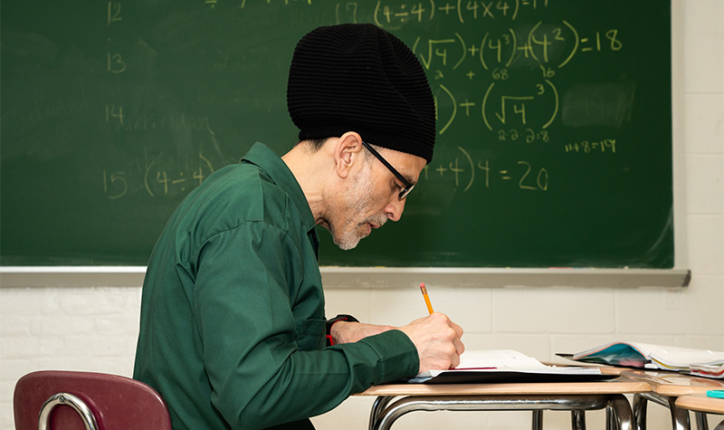 A BPI student writes in a notebook nearby a blackboard covered in equations.