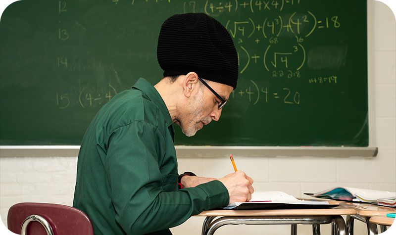 A student writes in a notebook by a blackboard covered in equations.